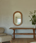 An Oak Oval Mirror with a solid wood frame hangs above a wooden console table decorated with a vase of leafy branches, reflecting a curtained window and outdoor greenery. A beige chair is partly visible against the light wall.