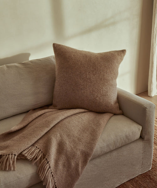 A beige sofa with the Alpaca Basketweave Pillow and a matching fringed blanket in a softly lit room with neutral walls.