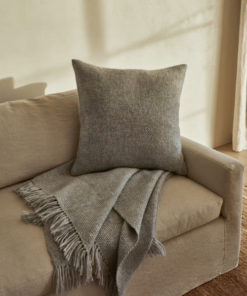 A beige sofa features the Alpaca Basketweave Pillow and a matching fringed gray throw blanket draped over the armrest, set against a light-colored wall with soft sunlight.