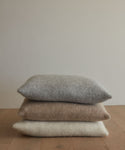 Three textured, knitted throw pillows—including the super-soft Alpaca Basketweave Pillow in gray, beige, and off-white—are stacked on a wooden floor against a plain light-colored wall.