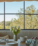 A dining table set with stacked plates, bowls, Roli Poli Glass smoke blue tumblers, and a vase of white tulips sits by a large window with trees and clear sky outside. Apples are placed near the bowls on the wooden table.