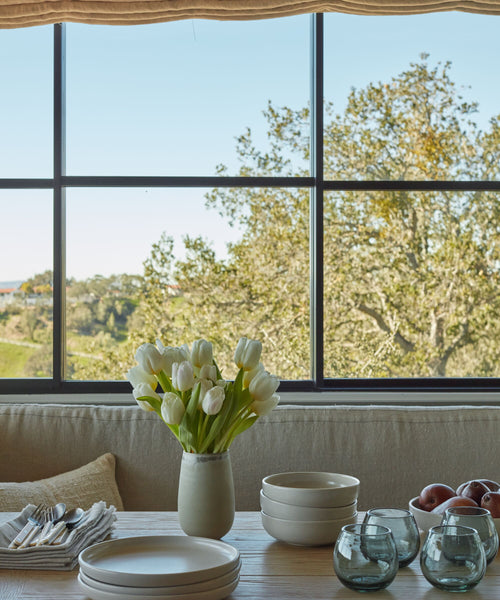 A dining table set with stacked plates, bowls, Roli Poli Glass smoke blue tumblers, and a vase of white tulips sits by a large window with trees and clear sky outside. Apples are placed near the bowls on the wooden table.