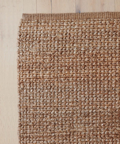 A close-up of the Bramble Rug, a handmade jute piece with a textured braided pattern, displayed on a light wooden floor. The warm brown natural rug is shown on the left side of the image.