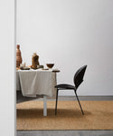 A minimalist dining area features a table with a beige tablecloth, ceramic dishes and vases, a black chair, and the Bramble Rug on a concrete floor against a plain white wall.