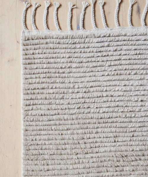 Close-up of the Malawi Rug, a hand-knotted, off-white wool piece with textured horizontal lines and fringe tassels along the top edge, handmade in India and displayed on a light wooden floor.