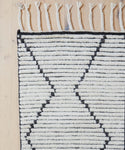 A close-up of the Nala Rug, a hand-knotted natural wool piece featuring black geometric zigzag lines and fringed tassels along the top edge, displayed on a light wooden floor.