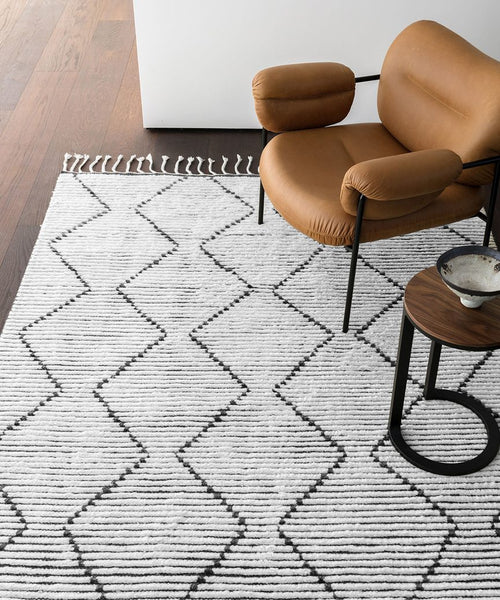 A modern tan armchair and small round side table rest on the Nala Rug, featuring a natural wool base with a black zigzag pattern and fringe, set atop a wooden floor near a white wall.