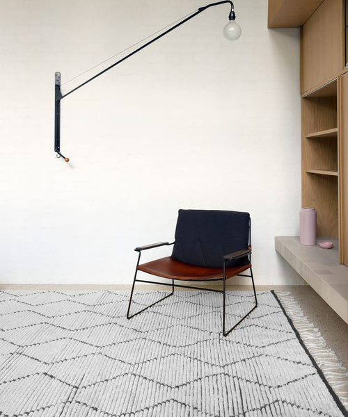 A minimalist room features a modern chair with a black leather seat, the hand-knotted Nala Rug in natural wool, built-in wooden shelves, a pink vase, and a long-arm wall lamp with an exposed bulb.