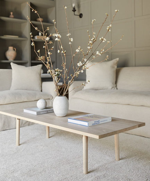 A minimalist living room with two beige sofas, a light wood coffee table topped with books and a decorative sphere, and blossoming branches in a vase. The Palermo Rug adds sustainable elegance to the calm atmosphere.