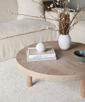 A round wooden coffee table with books, a white vase of blooming branches, a perforated white sphere, and a textured bowl sits before a beige sofa on the Palermo Rug in soft cream tones.