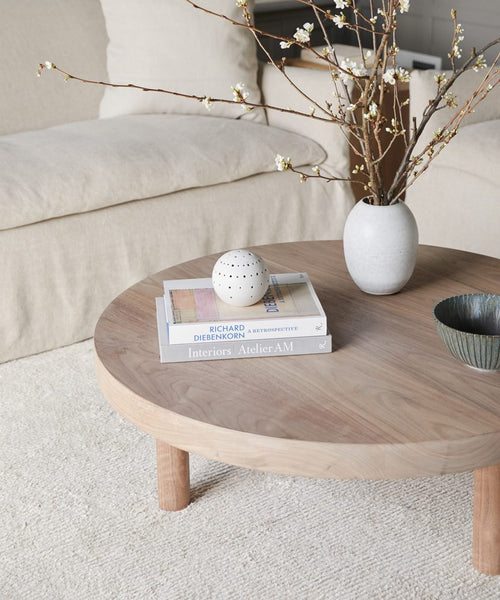 A round wooden coffee table with books, a white vase of blooming branches, a perforated white sphere, and a textured bowl sits before a beige sofa on the Palermo Rug in soft cream tones.