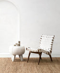 A modern woven white accent chair with wooden legs sits beside a small round white side table on the River Rug, set against a minimalist white wall with an arched niche.