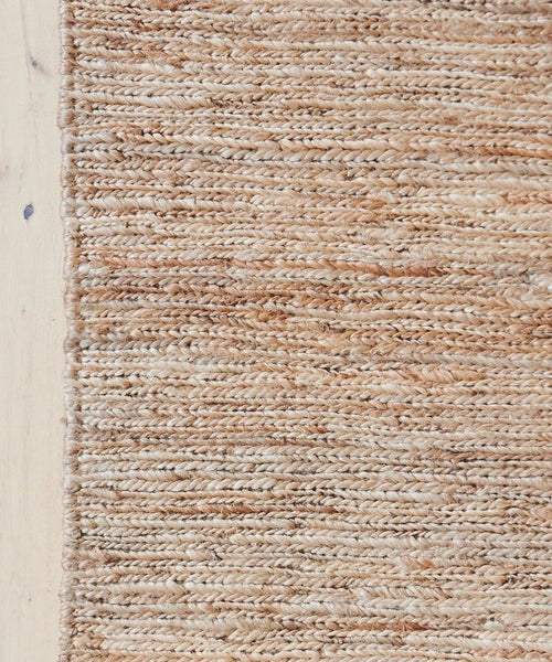 A close-up of the River Rug reveals its tan and beige jute fibers and textured braided patterns. The handwoven flatweave rug’s edge lies beside a light wooden floor, showcasing its artisanal craftsmanship.