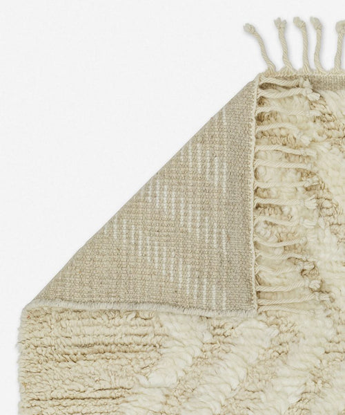The Cabin Rug from Jenni Kayne Rugs is a cream-colored, textured piece with a geometric pattern, flipped corner showing its woven underside and fringe—a Moroccan-inspired rug that adds timeless elegance to any space.