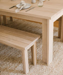 A light wood dining table and the Dining Bench sit on a woven rug. The table is simply set with a plate, napkin, fork, knife, and glass for an elegant minimalist dining space featuring handmade furniture.