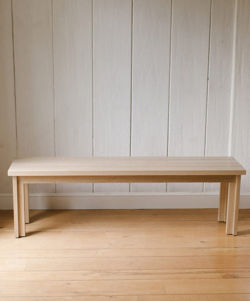 The Dining Bench, crafted from solid oak with straight legs, sits on a wooden floor against a white paneled wall.