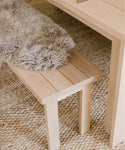 A Dining Bench made of light solid oak with a fluffy gray faux fur throw rests on a textured woven rug beside a matching wooden table.