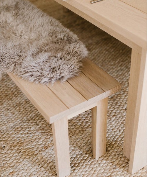 A Dining Bench made of light solid oak with a fluffy gray faux fur throw rests on a textured woven rug beside a matching wooden table.