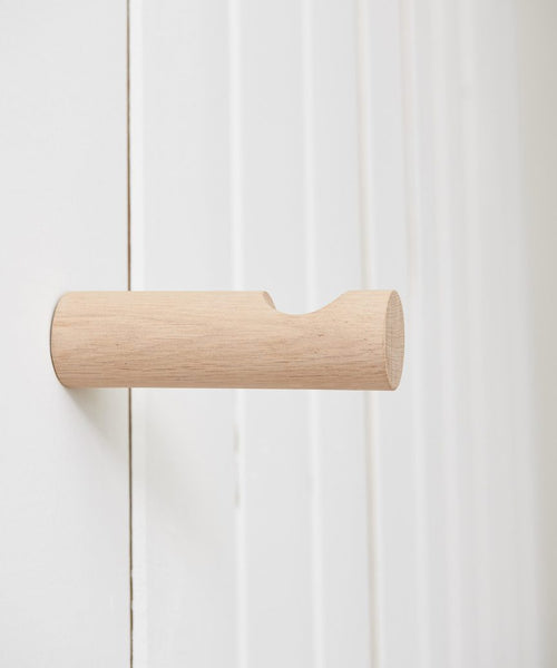 The Oak Wall Hook features a minimalist handmade rounded design in rustic white oak, shown mounted on a white wall with vertical paneling.