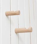 Two Oak Wall Hooks in rustic white oak are evenly spaced on a white vertical-paneled wall. Each cylindrical hook has a small notch near the end for hanging items.