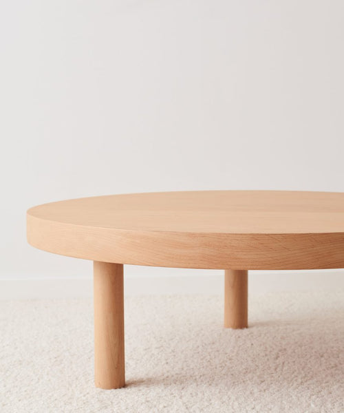 The Bay Coffee Table, crafted from knotty alder, showcases a minimalist round form with three cylindrical legs and is displayed on a light textured carpet in front of a plain white wall.