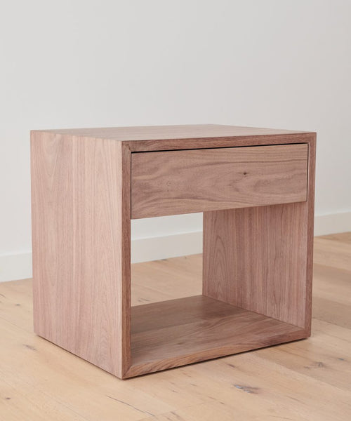 The Berkeley Nightstand is a modern, minimalist piece made of solid Walnut, featuring one drawer and an open shelf below. It’s shown on a light wood floor with a plain white wall background.