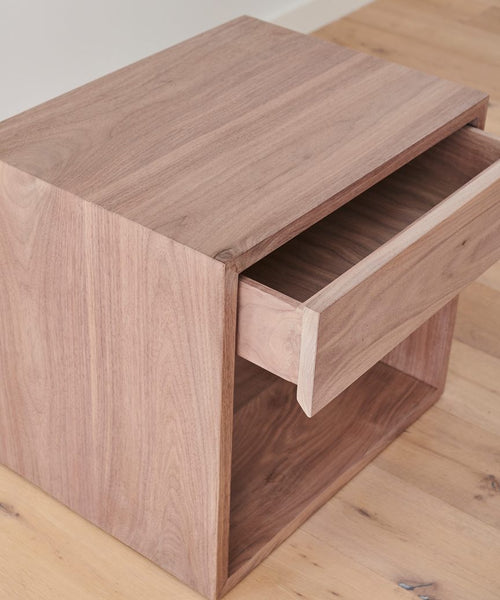 The Berkeley Nightstand is a minimalist, modern piece crafted from solid Walnut, featuring a handmade design with one open drawer and a shelf below—ideal for any contemporary space.