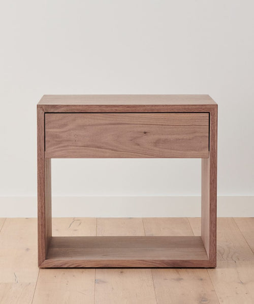 The Berkeley Nightstand features a minimalist, open-frame design in solid Walnut with a single drawer and bottom shelf, shown against a white wall and light wood floor.