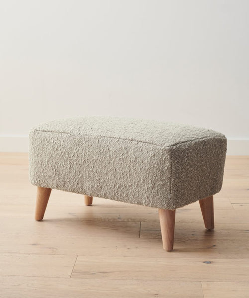 The Brentwood Ottoman features textured wool bouclé upholstery in light beige and four wooden legs, positioned on a light wood floor against a plain white wall.