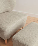 A close-up of a beige armchair in Belgian wool with wooden legs, paired with the Brentwood Ottoman, on a light wood floor against a plain white wall.