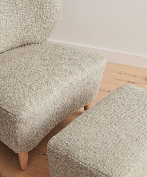 A close-up of a beige armchair in Belgian wool with wooden legs, paired with the Brentwood Ottoman, on a light wood floor against a plain white wall.