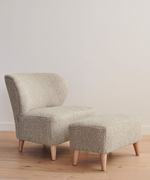 A modern accent chair in light beige Wool Boucle with wooden legs is paired with the Brentwood Ottoman, also on wooden legs, set on a wooden floor against a plain, light wall.