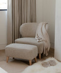 A cozy beige armchair with the Brentwood Ottoman, both topped with a soft throw blanket, sits by a window framed by floor-length curtains. Nearby on the wooden floor are fluffy slippers and a white rug.