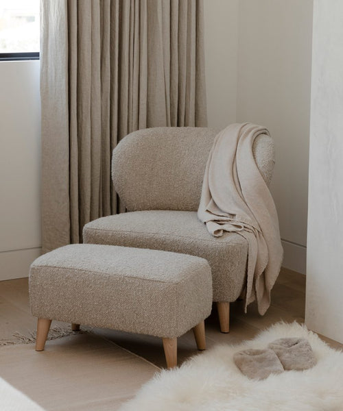 A cozy beige armchair with the Brentwood Ottoman, both topped with a soft throw blanket, sits by a window framed by floor-length curtains. Nearby on the wooden floor are fluffy slippers and a white rug.