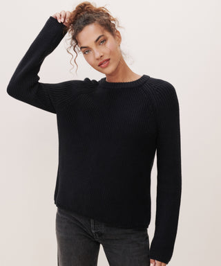 A person with curly hair wears a relaxed-fit black Cotton Fisherman Sweater and dark jeans, standing against a plain light background, touching their hair with a calm, relaxed expression.