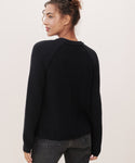 A woman with curly hair in a bun models the Cotton Fisherman Sweater and dark jeans, standing with her back to the camera and turning her head to the side against a plain light background.