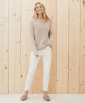 A smiling woman with long, loose hair stands before a light wooden wall, wearing a beige Cotton Fisherman Sweater, white pants, and beige slippers. One hand is in her pocket.