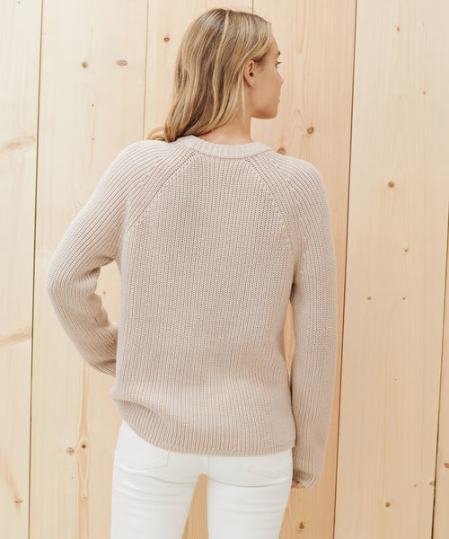 A woman with blonde hair, seen from the back, wears the Cotton Fisherman Sweater in light beige and white pants, standing before a light wooden wall.