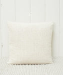 The Luna Pillow, a handwoven ivory square with a feather down insert, rests against a white wooden wall on a textured light rug.