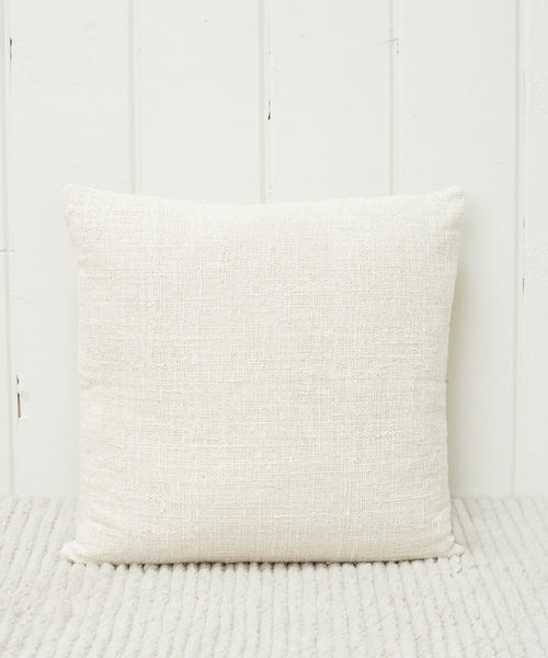 The Luna Pillow, a handwoven ivory square with a feather down insert, rests against a white wooden wall on a textured light rug.