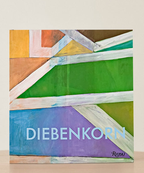 The book Richard Diebenkorn: A Retrospective, featuring a vibrant abstract geometric cover, stands upright on a wooden surface against a plain light background, celebrating the acclaimed California artist.