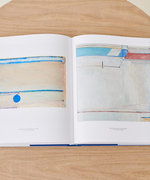 Richard Diebenkorn: A Retrospective is open on a light wooden table, displaying two abstract works with pastel colors, lines, and a striking blue circle on the left page by California artist Richard Diebenkorn.