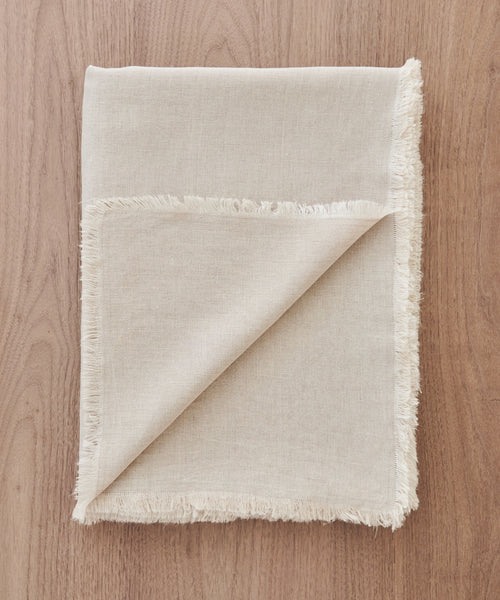 A beige, square 100% linen Frayed Linen Tablecloth with frayed edges rests on a light wooden surface, one corner turned up to reveal the underside—perfect for adding natural elegance to your dining space.