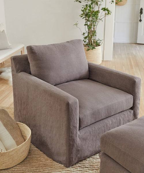 A handmade Harbor Chair with charcoal linen upholstery and a matching cushion is featured in a bright, minimalist living room with light wood floors, potted plants, and a woven rug.