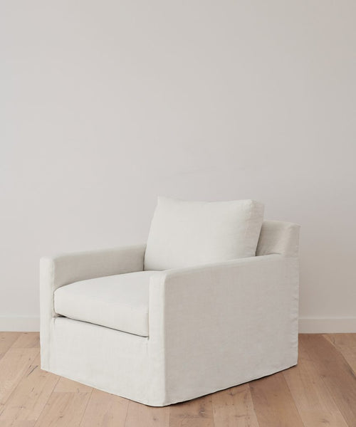 The Harbor Chair, featuring ivory linen upholstery and a boxy silhouette, stands on a light wood floor before a white wall, exemplifying the refined signature style of Jenni Kayne Furniture.