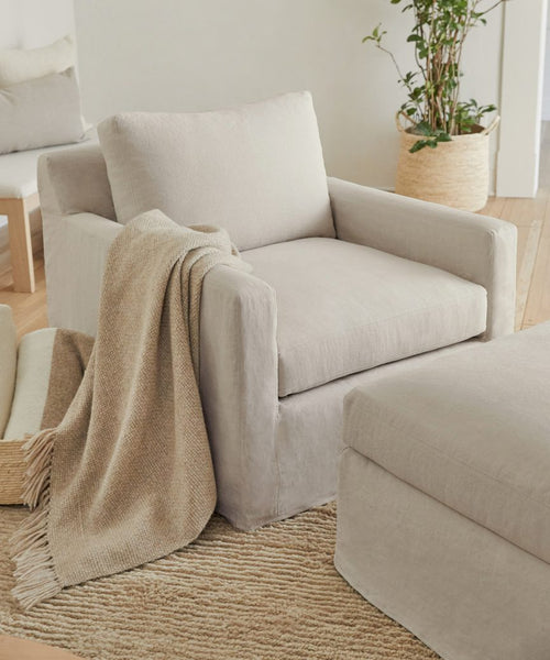 A cozy Harbor Chair in natural linen is paired with a soft, fringed tan throw and matching ottoman on a woven rug. Handmade in Los Angeles, it sits by a potted plant in a bright, minimal living space.