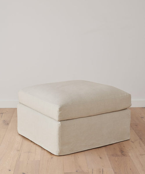The Harbor Ottoman, a handmade square piece in light beige natural linen, features a soft cushion and rests on a wooden floor against a plain white wall.