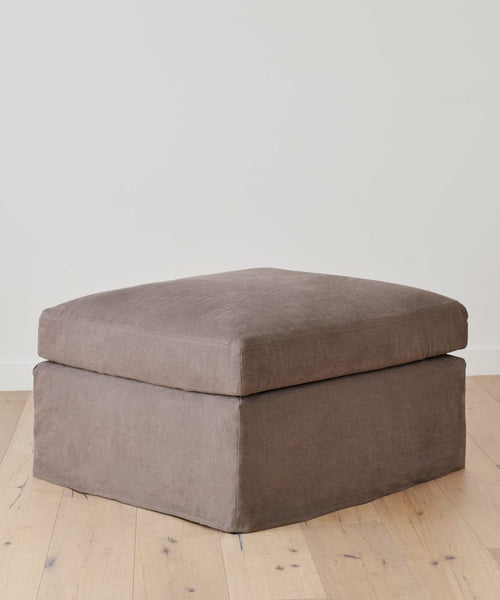 A taupe square Harbor Ottoman from Jenni Kayne Furniture, featuring a soft cushioned top, is placed on a light wood floor against a simple white wall.