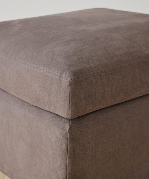 A close-up of the Harbor Ottoman in charcoal linen highlights its visible stitching and soft, cushioned top.
