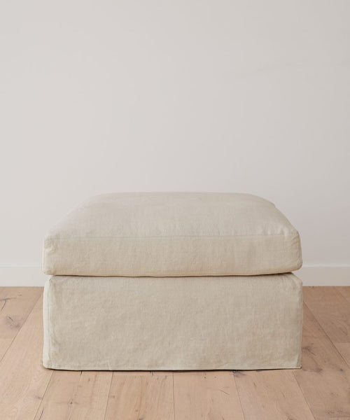 The Harbor Ottoman, handmade in Los Angeles and upholstered in natural linen with a soft cushion, rests on a light wooden floor against a plain white wall.